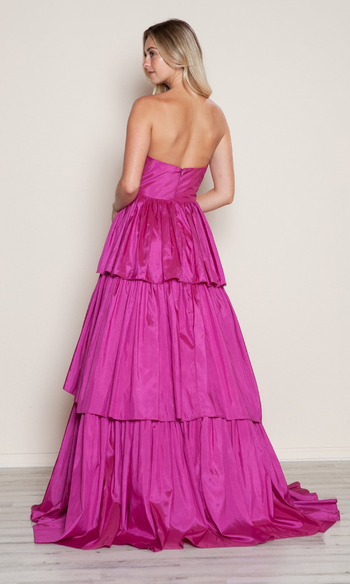 A woman stands with her back to the camera in the Long Prom Dress: Poly USA 9694—a bright pink, strapless, tiered A-line gown with a floor-length skirt.
